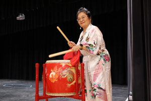 A performer plays a drum. She is wearing traditional Asian clothing