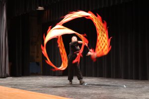 A performer on stage wears all black. The background is black. The performer is holding a red stick. Attached is a red and yellow paper dragon.