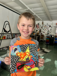 A young student stands in a classroom, holding a colorful piece of art. The student is smiling