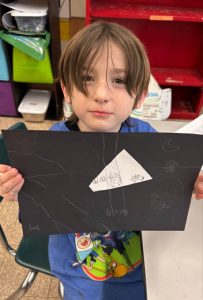 A young student stands in a classroom, holding a piece of artwork he created