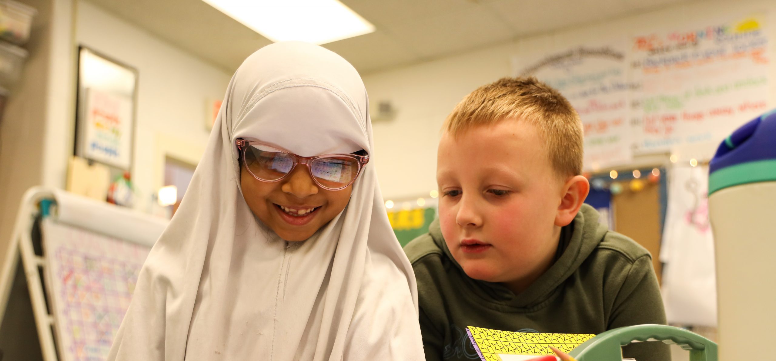 Two students doing classwork. One is holding a tablet and smiling and the other student is looking at the tablet.