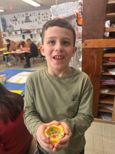 A young student stands in a classroom, holding a piece of artwork he created