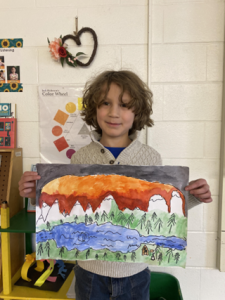 A young student stands in a classroom, holding a piece of colorful artwork he created