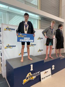 Three student-athletes stand on a tiered podium. The student standing at the first place spot holds a sign that says NYSPHSAA 2025 Champion and also wears a medal