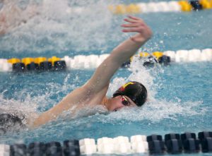 A swimmer is mid-stroke, swimming in a pool. The water is marked off by lane markers that are black and white and yellow and white. The swimmer wears googles and a black swim cap